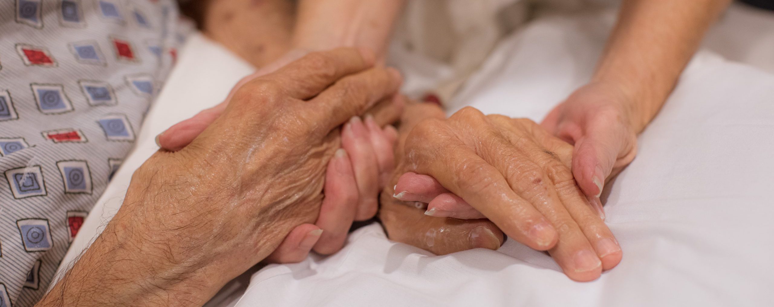 A close-up of an elderly person’s hands being gently held by a caregiver’s hands, symbolizing compassion, care, and support. The elderly person is wearing a patterned garment, and both are resting on a white bed.