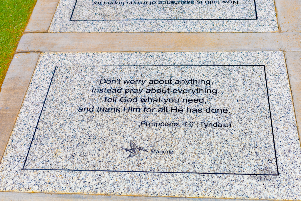 A granite plaque inscribed with the Bible verse Philippians 4:6 (Tyndale), which reads: 'Don't worry about anything. Instead, pray about everything. Tell God what you need and thank Him for all He has done.' The plaque also features an engraved hummingbird and the name Marjorie.