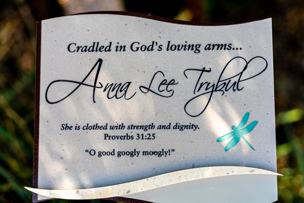 A close-up of a memorial plaque dedicated to Anna Lee Tribul, with the inscription 'Cradled in God's loving arms... Anna Lee Tribul. She is clothed with strength and dignity. Proverbs 31:25. "O good googly moogly!"' The plaque features a decorative design with a blue dragonfly.