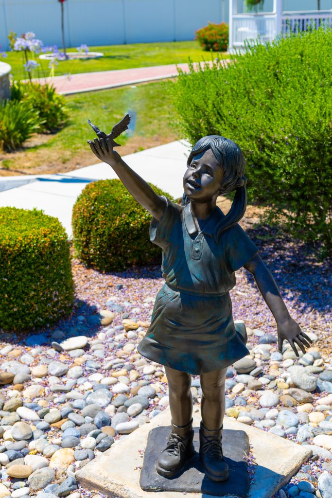 A bronze statue of a young girl joyfully reaching out her hand as a butterfly rests on her fingers. The statue is set in a beautifully landscaped garden with shrubs, flowers, and a stone pathway in the background.
