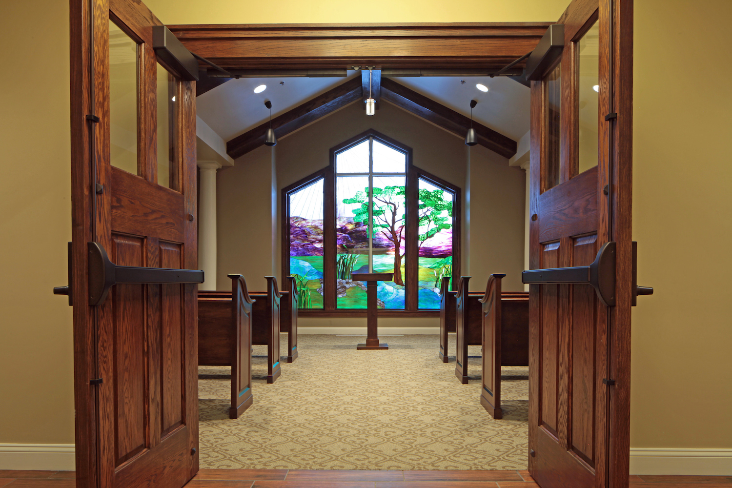 An open wooden door leading into a serene chapel with a vibrant stained glass window depicting a tree and nature scene. The room features wooden pews and a welcoming, peaceful atmosphere